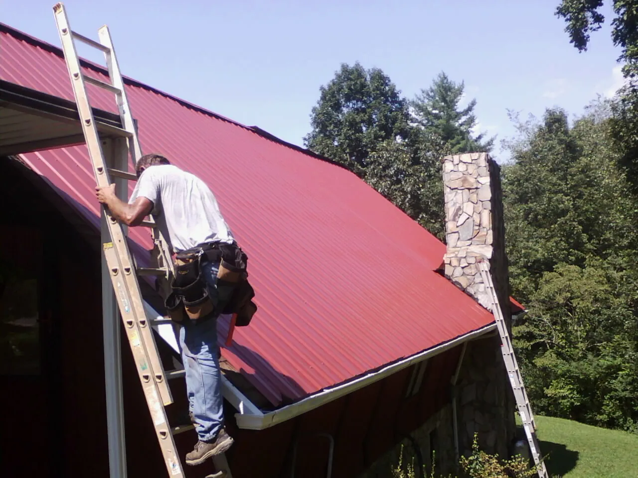 Roofer on a ladder beside a red metal roof and stone chimney — Soffit & Fascia Repair by Roofing Brothers in Sumner